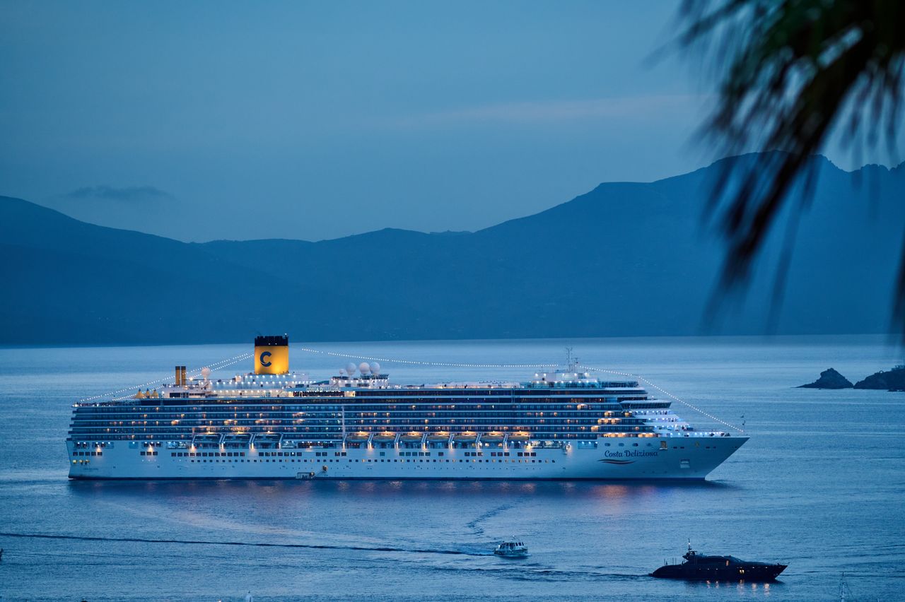 Services-01 A beautifully illuminated cruise ship sails across the calm sea at dusk, framed by mountains.